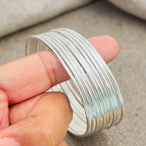 Può includere: Una serie di dieci braccialetti sottili e lisci in metallo color argento. I bracciali circolari hanno uno spessore uniforme e una finitura lucida e riflettente. Il set di braccialetti è tenuto insieme come un'unica unità, mostrando il riflesso.