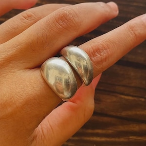 Puede incluir: Dos anillos de tono plateado con una superficie lisa, pulida y redondeada. Cada anillo presenta un diseño de banda gruesa y abovedada que se asienta de forma prominente en el dedo. El metal tiene un acabado reflectante que capta la luz.