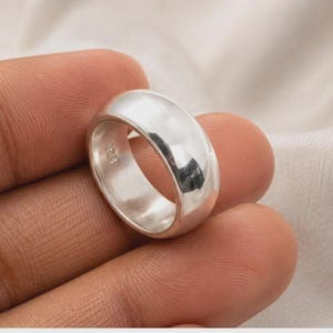 May include: A polished silver band ring with a smooth, rounded exterior surface. The interior of the band features a clear 925 hallmark stamp, indicating the metal composition. The ring has a simple, solid construction with a reflective finish.