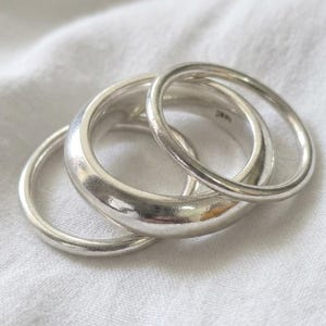 Puede incluir: Un conjunto de tres anillos de plata pulida de diferentes grosores. Los anillos están apilados, con bandas lisas y redondeadas que presentan un acabado metálico reflectante. El diseño consta de una banda gruesa y dos bandas más finas y delicadas.