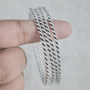 May include: A set of three silver-colored bangles, each featuring a twisted rope-like texture. The bangles are circular in shape and have a polished, metallic finish that reflects light along the ridges of the twisted metal design.