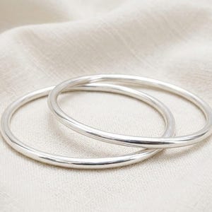Puede incluir: Dos pulseras tipo brazalete de plata pulida con un perfil liso y redondeado. Los brazaletes tienen forma circular y presentan un acabado metálico reflectante que capta la luz, mostrando un diseño de banda continua y sencilla sin adornos añadidos.
