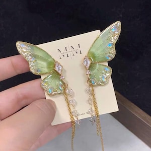May include: A pair of butterfly wing earrings featuring translucent light green resin with gold glitter accents and clear crystals. The earrings are attached to a cream-colored display card with the text MM MERRYMAKERS and Silver 925.