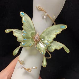 May include: A gold-toned bracelet featuring a large, translucent light green butterfly with iridescent accents and a central crystal embellishment. The band is a thin gold wire adorned with small white faux pearls and tiny white flower-shaped charms.