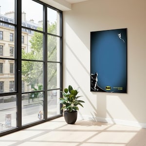 Puede incluir: Un póster enmarcado de Batman cuelga de una pared beige en una habitación con grandes ventanales. El póster presenta a Batman, una señal de murciélago y el texto "One with them, Las Vegas". Una planta en maceta está en el suelo.