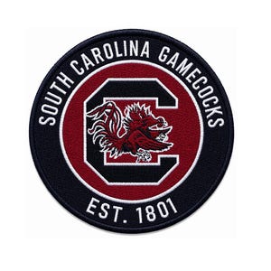 Puede incluir: Un parche circular con el logotipo de los Gamecocks de Carolina del Sur. El diseño presenta un gallo estilizado dentro de una 'C' granate sobre fondo negro, rodeado por el texto 'SOUTH CAROLINA GAMECOCKS' y 'EST. 1801'.