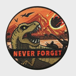 May include: Embroidered patch featuring a Tyrannosaurus Rex with an open mouth against a fiery sunset backdrop. The patch includes the text "NEVER FORGET" in orange letters. The design incorporates silhouettes of dinosaurs and flying creatures.