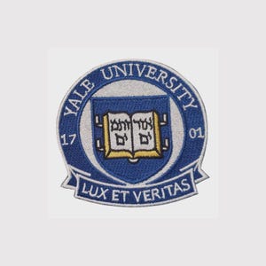 Pode incluir: Um emblema bordado circular com o selo da Universidade de Yale em azul, branco e dourado. O design inclui um livro aberto com texto em hebraico, as palavras Yale University, os números 17 e 01, e o lema latino Lux Et Veritas.