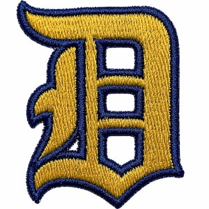 May include: Embroidered patch featuring a gold letter "D" with a navy blue outline. The letter has a classic, collegiate-style design, with three rectangular cutouts. The patch is likely for apparel or accessories.