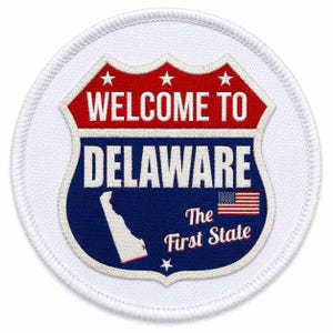 Puede incluir: Parche redondo blanco con un diseño de escudo rojo, blanco y azul. El escudo dice "WELCOME TO DELAWARE" y "The First State" con una pequeña bandera americana. Borde blanco.