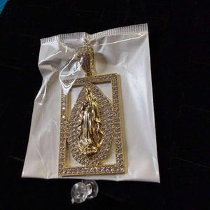 Puede incluir: Un colgante rectangular de tono dorado con un relieve ovalado central de la Virgen María, rodeado por un borde de pequeñas piedras claras. El colgante incluye una anilla grande incrustada con piedras en la parte superior y está en una bolsa.