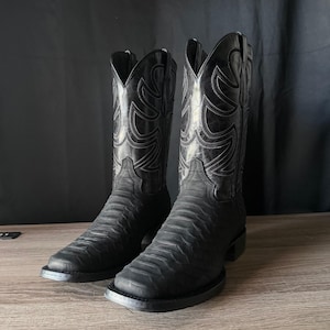 May include: A pair of black cowboy boots featuring a textured, scale-like pattern on the vamp and a smooth leather shaft with decorative tonal stitching. The boots have a square toe design, a stacked heel, and pull tabs at the top of the shaft.