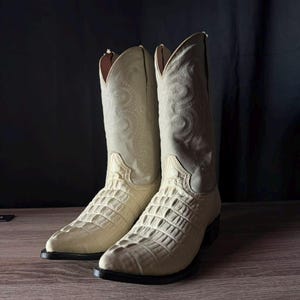 Könnte beinhalten: Ein Paar cremeweiße Cowboystiefel mit einem strukturierten, reptilienartigen Ledervorderblatt und einem hohen Schaft mit aufwendigen, farblich abgestimmten Ziernähten. Die Stiefel haben eine spitz zulaufende Kappe, einen Absatz und eine dunkle Sohle.