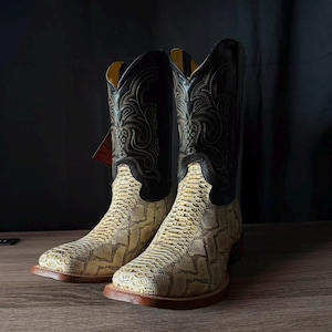May include: A pair of cowboy boots featuring a cream-coloured, textured snakeskin-patterned vamp and a black leather shaft. The black shaft is decorated with intricate, tonal stitching in a swirling pattern, and the boots have a square toe design.