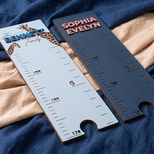 May include: Two rectangular wooden height charts with measurement markings. The light blue chart features giraffe illustrations and the text "The BENNETT Family". The dark blue chart features the raised pink text "SOPHIA EVELYN". Both include cutouts at the bottom.