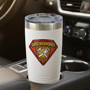 May include: A white insulated travel mug with a silver rim and a clear lid. The mug has a red, brown, and yellow diamond-shaped sticker with the words "SUPER WOODWORKER" and images of a hammer and saw.