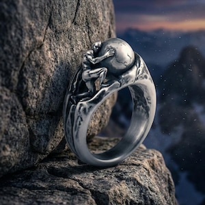 Puede incluir: Un anillo de plata con una figura esculpida abrazando un globo terráqueo. El diseño del anillo incluye una representación detallada de la figura y el globo, sobre un fondo rocoso texturizado. El anillo es una pieza de joyería única.