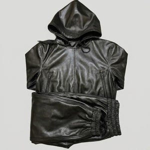 May include: A black leather hooded jacket and matching pants set. The jacket has a zipper closure and two front pockets. The pants have a gathered waistband. The set is made of a shiny, smooth leather material.