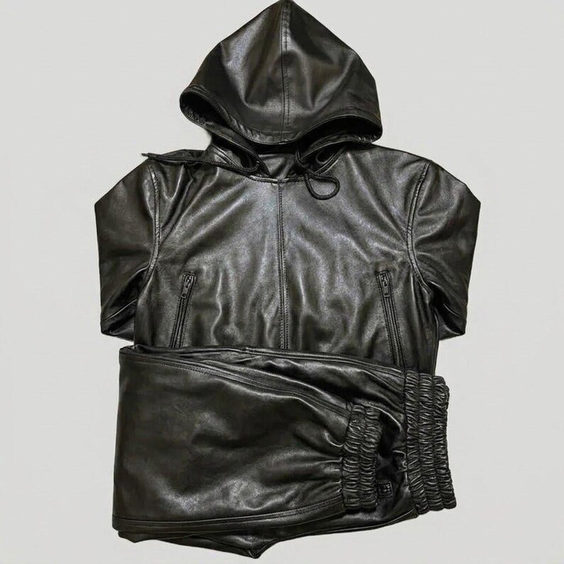 May include: A black leather hooded jacket and matching pants set. The jacket has a zipper closure and two front pockets. The pants have a gathered waistband. The set is made of a shiny, smooth leather material.