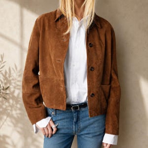 May include: A brown suede jacket with a classic collar and front button fastening, worn over a crisp white button-down shirt and blue denim jeans with a black belt. The jacket features long sleeves and a straight hemline, providing a structured look.
