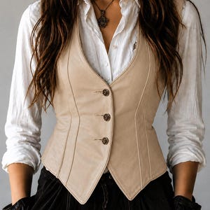 May include: A beige leather vest with three decorative buttons. The vest is worn over a white button-down shirt and black pants. The vest has a tailored fit and a V-neck design. The image shows a close-up of the vest and the upper body.