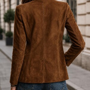 May include: A brown suede blazer featuring a structured collar, long sleeves, and a tailored fit. The jacket is worn with blue denim jeans. The back of the blazer displays a central vertical seam and clean, minimalist lines throughout the garment.