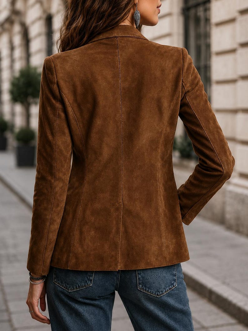 May include: A brown suede blazer featuring a structured collar, long sleeves, and a tailored fit. The jacket is worn with blue denim jeans. The back of the blazer displays a central vertical seam and clean, minimalist lines throughout the garment.