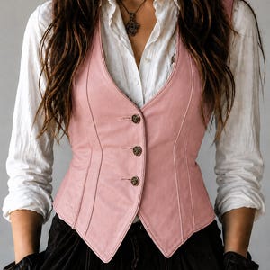 May include: A light pink leather vest with a V-neck and three decorative buttons. The vest is worn over a white button-down shirt. The model is also wearing black pants and a necklace.