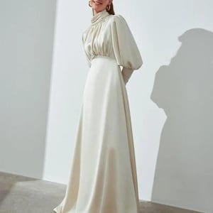 以下が含まれることがあります： クリーム色の、床まで届くロングドレス。ハイネックと長くてボリュームのある袖が特徴です。ドレスは体にフィットする胴と流れるようなスカートが特徴です。モデルは金のフープイヤリングをしています。ドレスは滑らかなサテンのような生地で作られています。