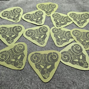 May include: Several olive green, triangular patches with a dark grey ram skull design. The patches are laid out on a grey fabric. The skull design includes curved horns and detailed facial features.