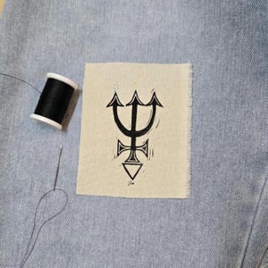 Peut inclure: Un écusson rectangulaire en tissu blanc cassé arborant un symbole de trident noir imprimé avec une base en forme de croix et de triangle. L'écusson est posé sur un tissu en jean bleu clair à côté d'une bobine de fil noir et d'une aiguille.
