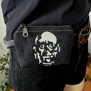 May include: A black denim pouch featuring a white graphic print of a stylised face. The pouch has a silver-toned metal zip closure with a circular pull tab and a small metal ring attached to the side, secured to a brown leather belt.