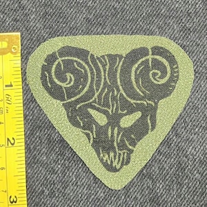 Peut inclure: Un écusson en tissu triangulaire vert olive clair avec un motif sérigraphié noir représentant un crâne de bélier aux cornes recourbées. Un mètre ruban jaune est placé à gauche, indiquant une largeur d'environ 6,5 cm pour l'écusson.