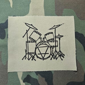 Könnte beinhalten: Ein quadratischer Aufnäher mit einer schwarzen Strichzeichnung eines Schlagzeugs. Das Schlagzeug umfasst eine Bassdrum, eine Snare-Drum, zwei Tomtoms und zwei Becken. Der Aufnäher befindet sich auf einem Tarnhintergrund.
