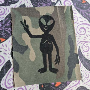 Peut inclure: Un écusson en tissu rectangulaire arborant un motif camouflage dans des tons vert olive, marron et beige. Au centre, une silhouette noire d'extraterrestre fait un signe de paix d'une main et se tient debout, les pieds légèrement écartés.