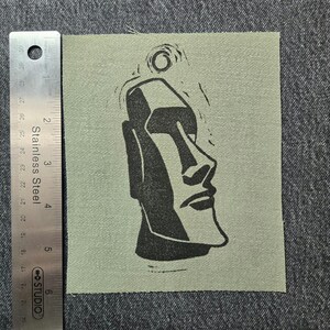 Peut inclure: Un écusson carré en tissu vert olive avec un dessin noir d'une tête Moai de l'île de Pâques. L'écusson mesure environ 10 cm de large. Une règle en acier inoxydable est placée à côté de l'écusson pour l'échelle.