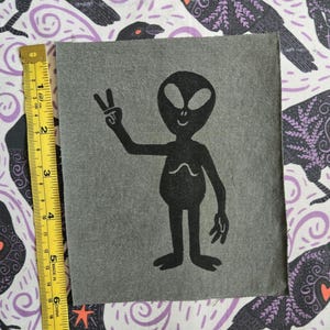 Könnte beinhalten: Ein grauer Aufnäher mit einer schwarzen Alien-Illustration, die ein Friedenszeichen zeigt. Der Alien hat große Augen und ein gebogenes Lächeln. Der Aufnäher ist etwa 12,7 cm hoch und 10,2 cm breit.