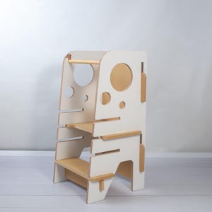 Puede incluir: Una torre de aprendizaje de madera clara y blanca con una plataforma y un escalón. Los paneles laterales tienen recortes circulares y ranuras horizontales. La estructura está construida con una combinación de paneles blancos y madera natural.