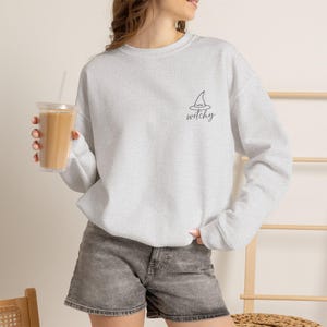 May include: A light gray crewneck sweatshirt featuring a small black line drawing of a witch hat on the upper left chest area, accompanied by the word "witchy" written in a cursive script font directly underneath the hat graphic.