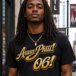 May include: A black short-sleeved t-shirt featuring the gold and white text Aaaa-Phiii! 06! printed across the chest in a stylized script font with a bold outline. The shirt has a crew neckline and is made of a solid black fabric material.