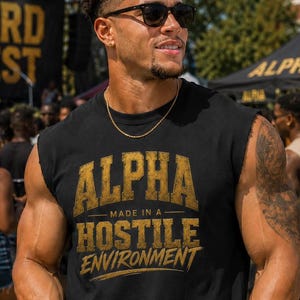 May include: A black sleeveless shirt featuring gold distressed text that reads ALPHA MADE IN A HOSTILE ENVIRONMENT. The shirt is paired with matching black shorts with the number 1906 printed in gold. A black cup with gold text is held in hand.