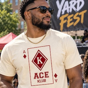 May include: A cream-colored short-sleeve t-shirt featuring a large graphic of a playing card on the chest. The graphic displays a diamond suit, the letter K inside a diamond, and the text ACE KLUB in a bold, dark red collegiate-style font.