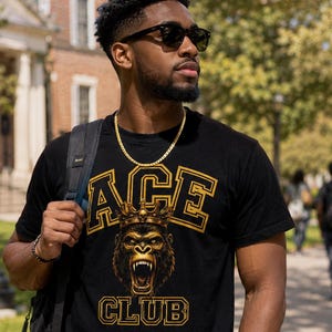 May include: A black short-sleeved t-shirt featuring a gold-colored graphic print of a gorilla wearing a crown. The text ACE is printed in a gold collegiate font above the gorilla, and the word CLUB is printed in the same font below the graphic.