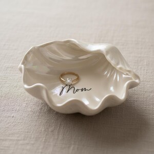 May include: A white, shell-shaped ring dish with a gold ring featuring a clear stone. The dish has the word "Mom" written in black script. The dish is pearlescent and sits on a neutral-colored surface.