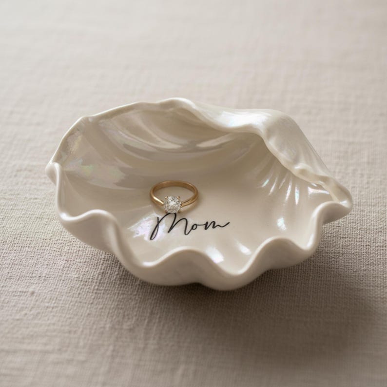 May include: A white, shell-shaped ring dish with a gold ring featuring a clear stone. The dish has the word "Mom" written in black script. The dish is pearlescent and sits on a neutral-colored surface.