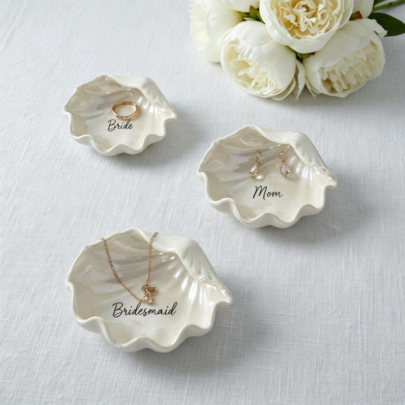 May include: Three off-white, shell-shaped trinket dishes. One holds a gold ring and says "Bride", another has earrings and says "Mom", and the third has a necklace and says "Bridesmaid". White flowers are in the background.