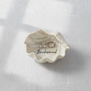 May include: A cream-colored, seashell-shaped trinket dish with the word "Bridesmaid" in black script. The dish holds a pair of stud earrings and a silver ring. The dish is on a white surface.