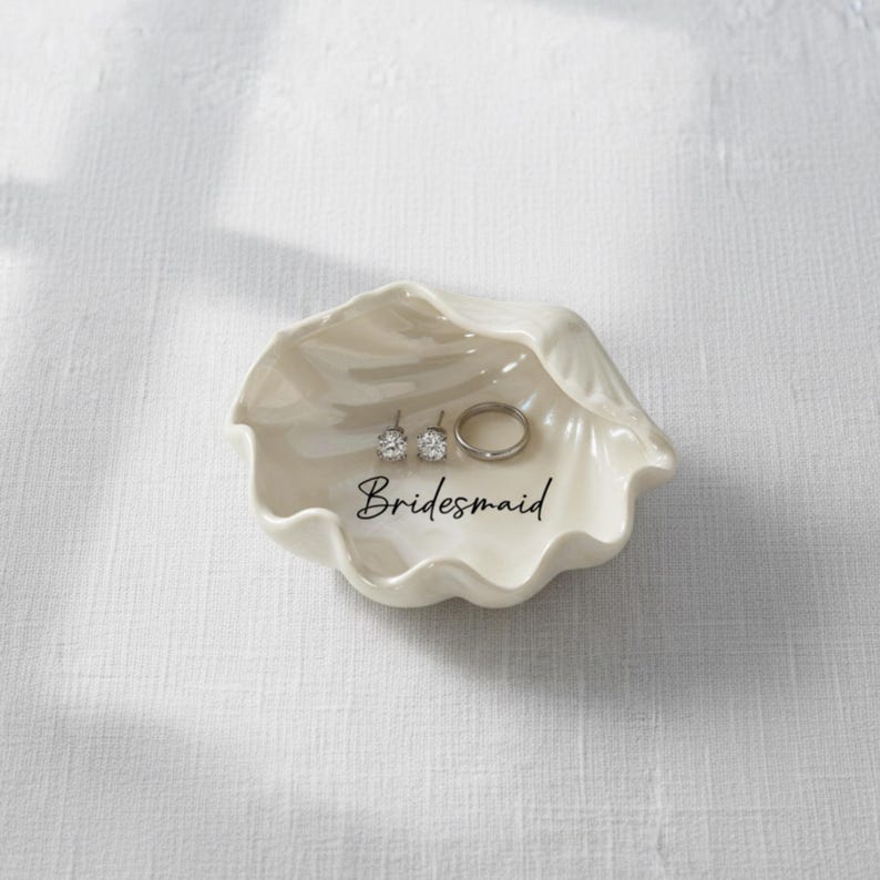 May include: A cream-colored, seashell-shaped trinket dish with the word "Bridesmaid" in black script. The dish holds a pair of stud earrings and a silver ring. The dish is on a white surface.