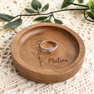 Pode incluir: Uma bandeja redonda de madeira para anéis com uma gravura floral e o nome "Martina". Um anel de diamante está no centro. A bandeja é marrom e tem cerca de 10 cm de diâmetro.