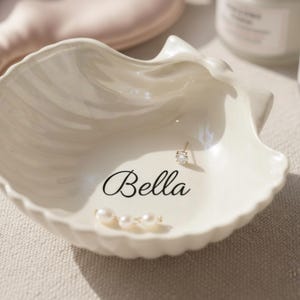 May include: A white, seashell-shaped trinket dish with the name "Bella" in cursive script. The dish holds a small, gold-colored earring with a clear gemstone and three small pearls. The dish is on a neutral-colored surface.